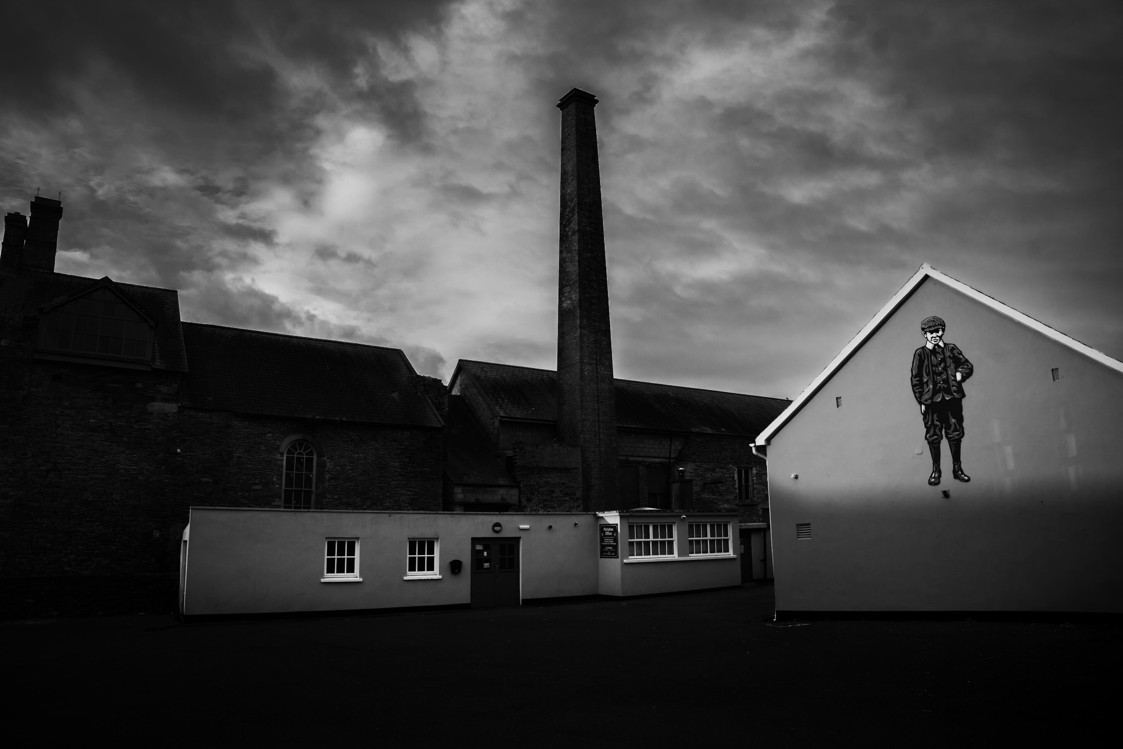 Black and white photograph of the Parish Centre in Clonakilty, West Cork, Ireland, featuring historic buildings beneath dramatic storm clouds and the iconic mural on the gable wall.
